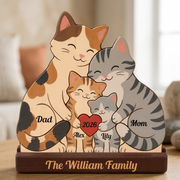 A personalized wooden family plaque shaped like a cat family, with a calico cat labeled Dad, a gray tabby cat labeled Mom, and two smaller orange and gray kittens names, all gathered around a red heart that says 2026. The base is engraved with 'The Family'.