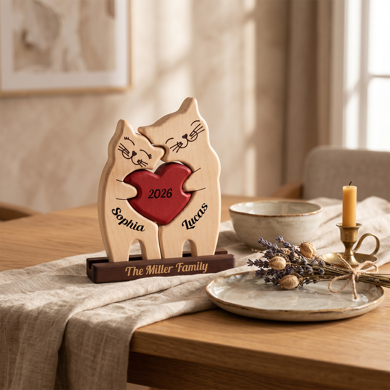 Personalized wooden cat figurines hugging with a red heart in the center, engraved with the names, The figurines are styled on a wooden dining table with a beige cloth, ceramic dishes, dried lavender, and a candle.