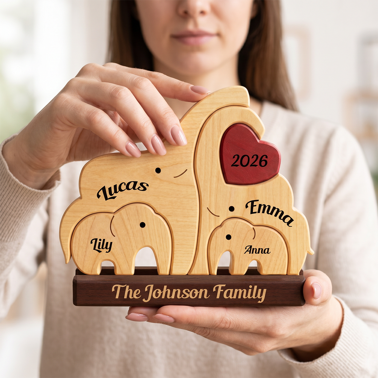 A woman holds a personalized wooden family elephant plaque. The set features four light natural wood elephants of varying sizes, each engraved with a child's name 