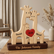 Personalized wooden giraffe family sculpture on a dark base, with three light wood giraffes and a red heart in the center reading '2026', plus the names  and the family name. The piece is photographed on a wooden table in a softly styled room with curtains, a chair, and a vase of dried grass.