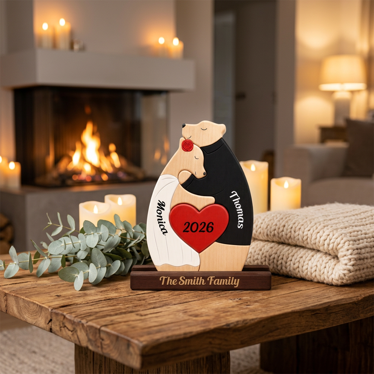 Personalized wooden polar bear couple figurine on a dark base, shown on a rustic table in a cozy living room with a fireplace, candles, and a knit blanket. The bears are dressed in white and black wedding attire with a red heart in the center reading '2026' and the names on the figures, and 'The Family' engraved on the base.