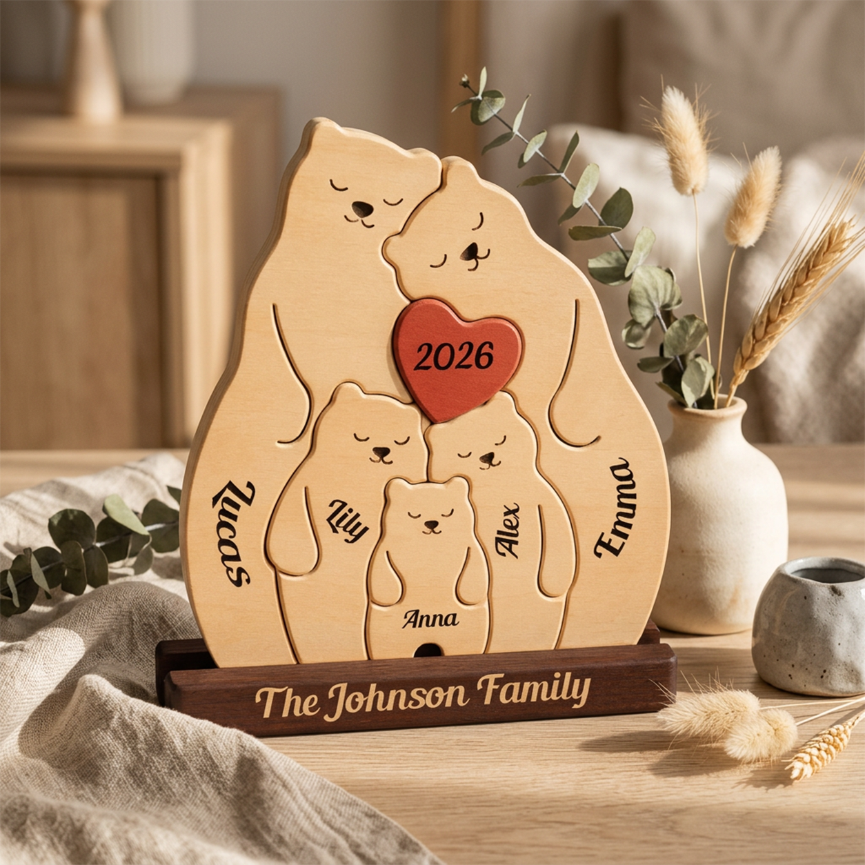 Personalized wooden family plaque shaped like a bear family, engraved with names and a red heart reading 2026, displayed on a wooden stand in a warm home setting with dried grasses and a small vase.