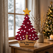 A red wooden Christmas tree cutout with white polka dots and a gold star topper sits on a wooden table in a cozy indoor setting. The base is engraved with 'Merry Christmas 2026'.
