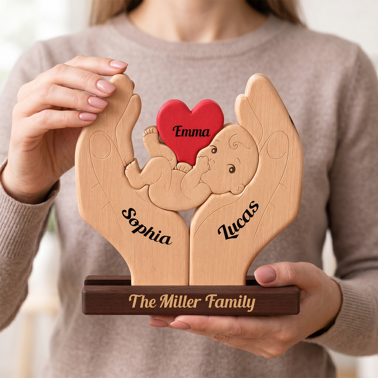 A person in a beige sweater holds a personalized wooden keepsake plaque shaped like two hands cradling a baby figure. 