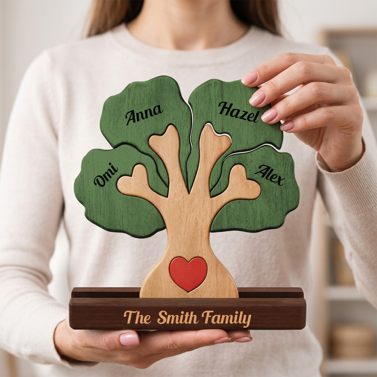 A personalized wooden family tree display held by a person. The tree has green leaves engraved with family names. Natural wood trunk, a red heart at the base, and a dark wood plaque engraved with 'The Family Name'.