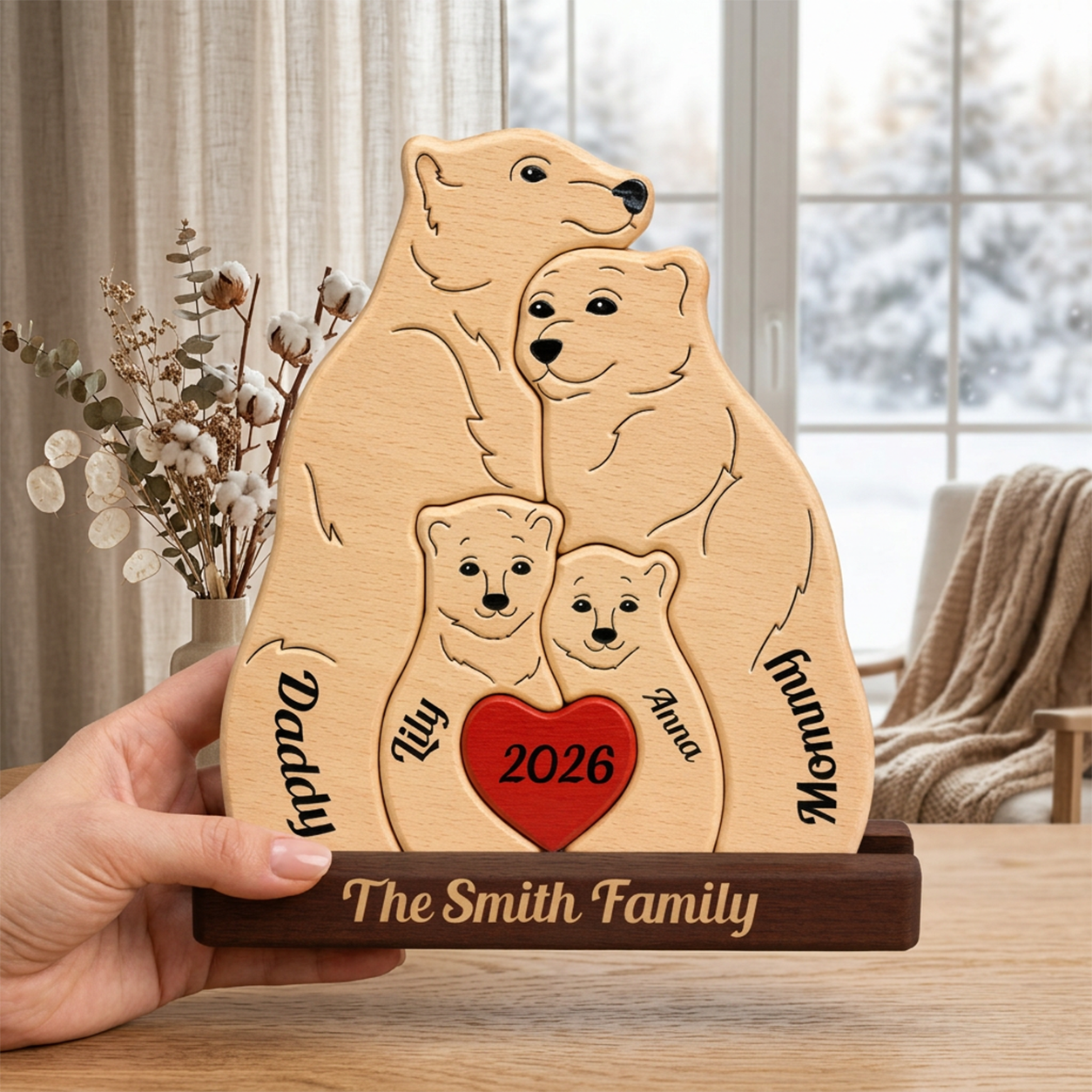 A personalized wooden family plaque shaped like a polar bear family, held by a hand in a home setting. The light natural wood plaque features engraved black line art of a polar bear with four cubicles, a red heart in the center with '2026', and custom names 